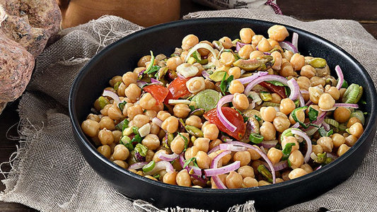 Kichererbsensalat mit Kirschtomaten, grünen Oliven, roten Zwiebeln und Pistazien in einer dunklen Schüssel.